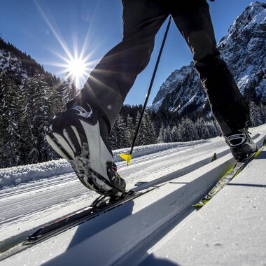 A skier glides over a snow-covered cross-country skiing trail. In the background, the mountains shine under a clear blue sky.