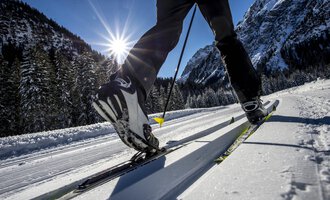 A skier glides over a snow-covered cross-country skiing trail. In the background, the mountains shine under a clear blue sky.