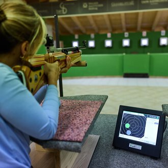 Ein Schütze zielt mit einem Gewehr auf eine Zielscheibe. Im Vordergrund ist ein Monitor sichtbar, der die Ergebnisse anzeigt. | © Kleinwalsertal Tourismus | Oliver Farys