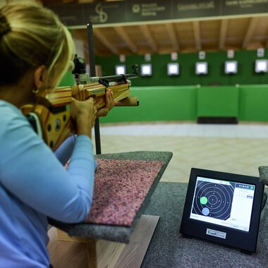 Ein Schütze zielt mit einem Gewehr auf eine Zielscheibe. Im Vordergrund ist ein Monitor sichtbar, der die Ergebnisse anzeigt. | © Kleinwalsertal Tourismus | Oliver Farys