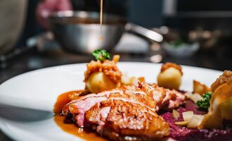 Ein köstliches Gericht auf einem weißen Teller, dekoriert mit Sauce und Beilagen. Im Hintergrund ist ein Koch zu sehen, der Sauce über das Essen träufelt. | © Genuss- & Aktivhotel Sonnenburg | Frank Simon
