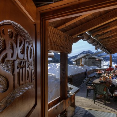 Ein gemütlicher Gasthof mit Holzverzierungen und Blick auf schneebedeckte Berge. Draußen sitzen Gäste an einem Tisch und genießen die alpine Atmosphäre. | © Bergstüble | Michaela Beck Dornach
