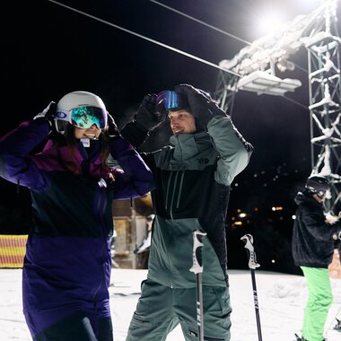 Ein Paar Skifahrer bereitet sich auf das Skifahren bei Nacht vor. Im Hintergrund sind weitere Skifahrer und eine beleuchtete Skiliftanlage zu sehen. | © Kleinwalsertal Tourismus | Oliver Farys