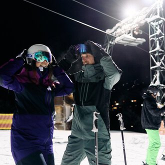 Ein Paar Skifahrer bereitet sich auf das Skifahren bei Nacht vor. Im Hintergrund sind weitere Skifahrer und eine beleuchtete Skiliftanlage zu sehen. | © Kleinwalsertal Tourismus | Oliver Farys