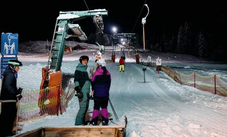 Skifahrer warten an einem beleuchteten Skilift in der Nacht. Die Pisten sind schneebedeckt und umgeben von Bäumen. | © Kleinwalsertal Tourismus | Oliver Farys