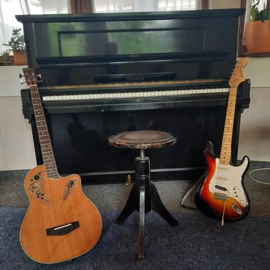Ein Klavier steht im Hintergrund, flankiert von einer akustischen Gitarre links und einer E-Gitarre rechts. In der Mitte befindet sich ein Hocker. | © Kulturcafe Kleinwalsertal | Gerhard Bauer