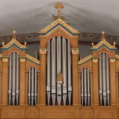 A magnificent organ with ornate decorations in warm wooden colors. The pipes are symmetrically arranged and rise above the stage area. | © Karl Kessler