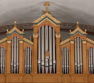 A magnificent organ with ornate decorations in warm wooden colors. The pipes are symmetrically arranged and rise above the stage area. | © Karl Kessler