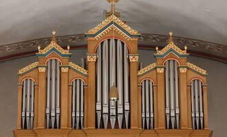 A magnificent organ with ornate decorations in warm wooden colors. The pipes are symmetrically arranged and rise above the stage area. | © Karl Kessler
