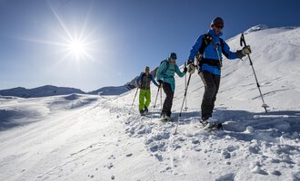 Eine Gruppe von Menschen wandert durch eine verschneite Landschaft. Die Sonne scheint klar am Himmel. | © Kleinwalsertal Tourismus eGen | Dominik Berchtold