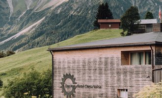 Ein modernes Gebäude in den Bergen mit einer Holzfassade. Die Umgebung ist grün mit einer beeindruckenden Berglandschaft im Hintergrund. | © Das Naturhotel Chesa Valisa
