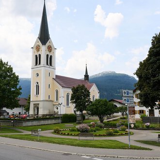 A picturesque church with a tall tower stands in the center of a small square. In the background, green trees and mountains can be seen.