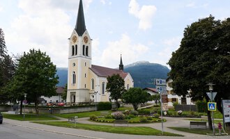 A picturesque church with a tall tower stands in the center of a small square. In the background, green trees and mountains can be seen.