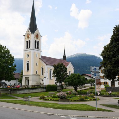 A picturesque church with a tall tower stands in the center of a small square. In the background, green trees and mountains can be seen.