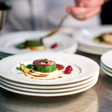 Ein aufmerksam angerichteter Teller mit feinem Essen. Im Hintergrund sieht man einen Koch, der eine Sauce zubereitet. | © Ifen Hotel Kleinwalsertal | Annette Sandner