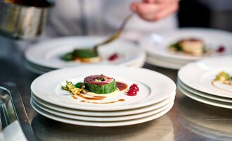 A carefully arranged plate with fine food. In the background, you can see a chef preparing a sauce. | © Ifen Hotel Kleinwalsertal | Annette Sandner