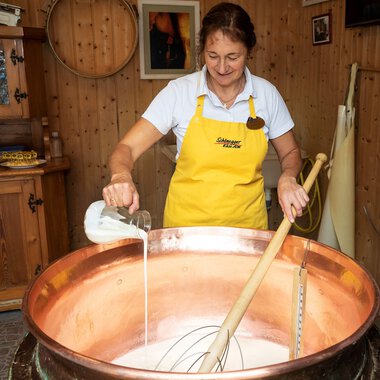 Eine Frau in einem gelben Schürze rührt in einem großen Kupfertopf mit Milch. Im Hintergrund sind Holzregale und eine gemütliche Einrichtung zu sehen. | © Schönegger Käsealm