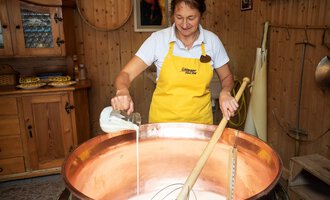Eine Frau in einem gelben Schürze rührt in einem großen Kupfertopf mit Milch. Im Hintergrund sind Holzregale und eine gemütliche Einrichtung zu sehen. | © Schönegger Käsealm