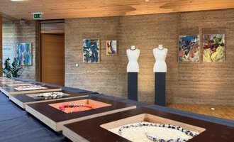Eine moderne Ausstellung mit mehreren Schmuckstücken in Vitrinen. Im Hintergrund stehen Schaufensterpuppen und bunte Gemälde an der Wand. | © Gerti Maitz