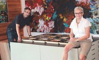 Ein Mann und eine Frau stehen an einem Tisch, auf dem zahlreiche Bilderrahmen liegen. Im Hintergrund sind farbenfrohe abstrakte Gemälde zu sehen. | © Gerti Maitz
