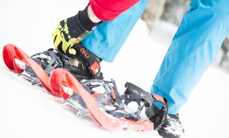 Eine Person schnallt Schneeschuhe an, während sie auf dem weißen Schnee steht. Der Hintergrund ist unscharf, was auf einen winterlichen Wald hinweist. | © Kleinwalsertal Tourismus | Oliver Farys