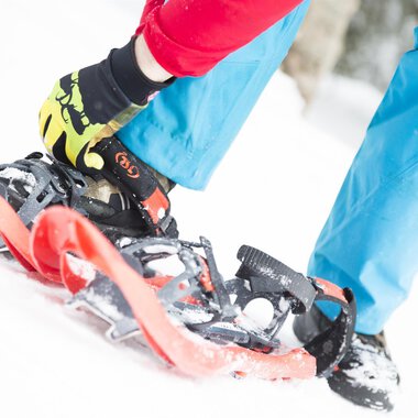 A person straps on snowshoes while standing in the snow. The ground is white and the clothing is colorful. | © Kleinwalsertal Tourismus eGen | Oliver Farys