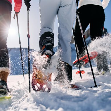 Drei Personen wandern im Schnee mit Schneeschuhen. Die Sonne scheint und Schnee spritzt von ihren Bewegungen auf. | © Kleinwalsertal Tourismus | Oliver Farys