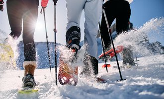 Drei Personen wandern im Schnee mit Schneeschuhen. Die Sonne scheint und Schnee spritzt von ihren Bewegungen auf. | © Kleinwalsertal Tourismus | Oliver Farys