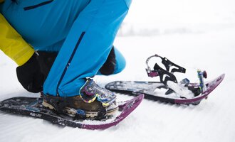 A person is putting on snowshoes while sitting on a snow-covered ground. The equipment and clothing are suitable for winter outdoor activities. | © Kleinwalsertal Tourismus | Oliver Farys