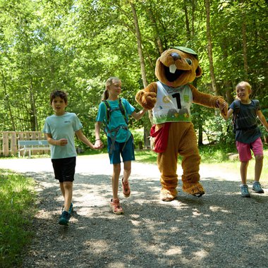 Kinder gehen fröhlich auf einem Weg durch den Wald, begleitet von einem lustigen Maskottchen. Die Atmosphäre ist freundlich und einladend. | © Kleinwalsertal Tourismus | Oliver Farys