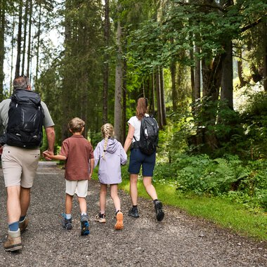 Eine Familie wandert auf einem Waldweg. Umgeben von hohen Bäumen und grüner Vegetation genießen sie die Natur. | © Kleinwalsertal Tourismus | Oliver Farys