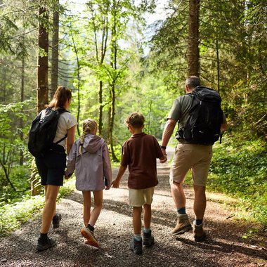 Eine Familie wandert gemeinsam auf einem Waldweg. Sie genießen die Natur um sich herum zwischen hohen Bäumen. | © Kleinwalsertal Tourismus | Oliver Farys