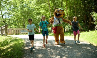 Eine fröhliche Gruppe von Kindern läuft auf einem Weg im Wald mit einem Maskottchen. Die Sonne scheint durch die Bäume und es herrscht eine entspannte Atmosphäre. | © Kleinwalsertal Tourismus | Oliver Farys