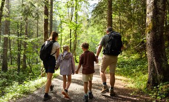 Eine Familie wandert gemeinsam auf einem Waldweg. Sie genießen die Natur um sich herum zwischen hohen Bäumen. | © Kleinwalsertal Tourismus | Oliver Farys