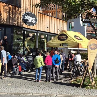 Eine Gruppe von Menschen steht vor einem Café mit einer gelben Markise. Es ist ein sonniger Tag, und einige Fahrer warten, während andere Radfahrer ihre Räder abstellen. | © Schönegger Käsealm