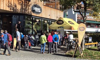 Eine Gruppe von Menschen steht vor einem Café mit einer gelben Markise. Es ist ein sonniger Tag, und einige Fahrer warten, während andere Radfahrer ihre Räder abstellen. | © Schönegger Käsealm
