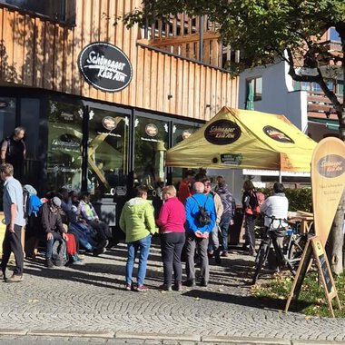 Eine Gruppe von Menschen steht vor einem Café mit einer gelben Markise. Es ist ein sonniger Tag, und einige Fahrer warten, während andere Radfahrer ihre Räder abstellen. | © Schönegger Käsealm