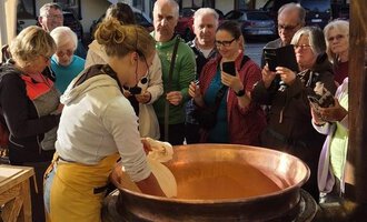 Eine Person bereitet etwas in einem großen Topf unter einem Zelt vor. Im Hintergrund schaut eine Gruppe von Menschen interessiert zu. | © Schönegger Käsealm