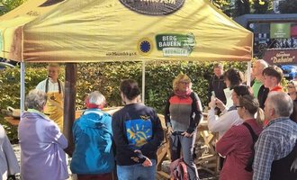 A sunny marketplace with a yellow sales booth. Many people are standing in a line waiting for products. | © Schönegger Käsealm