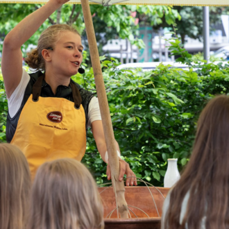 Eine Frau erklärt etwas mit einem langen Stock in der Hand. Im Hintergrund sind Kinder zu sehen, die interessiert zuhören. | © Schönegger Käsealm Susanne Lang