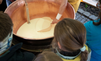 Eine Person rührt eine Mischung in einem großen Kupferkessel. Im Hintergrund sind interessierte Zuschauer, darunter Kinder, die neugierig zuschauen. | © Schönegger Käsealm Susanne Lang
