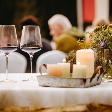 A beautifully set table with two wine glasses and rustic decor. In the background, guests can be seen at an event. | © Kleinwalsertal Tourismus eGen | Oliver Farys