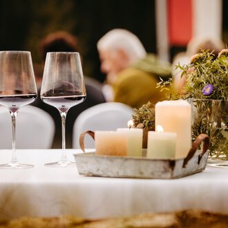 Ein schön gedeckter Tisch mit zwei Weingläsern und einer rustikalen Dekoration. Im Hintergrund sind Gäste auf einer Veranstaltung zu sehen. | © Kleinwalsertal Tourismus eGen | Oliver Farys