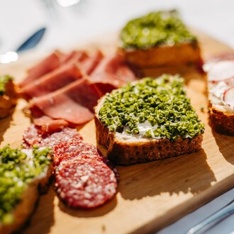A wooden board with various open-faced sandwiches. The snacks are garnished with herbs, sausage, and radishes. | © Kleinwalsertal Tourismus eGen | Oliver Farys