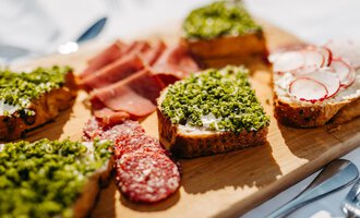 A wooden board with various open-faced sandwiches. The snacks are garnished with herbs, sausage, and radishes. | © Kleinwalsertal Tourismus eGen | Oliver Farys