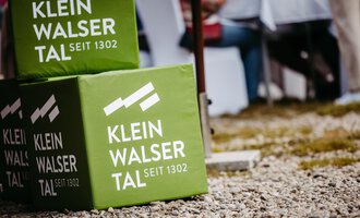 A green cube with the inscription "Klein Walsertal since 1302" stands on a gravel floor. Blurry tables and chairs can be seen in the background. | © Kleinwalsertal Tourismus eGen | Oliver Farys