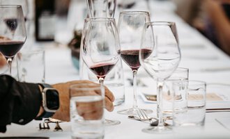 A table set with glasses for red wine and water. A hand holds a water glass while the background remains blurred. | © Kleinwalsertal Tourismus eGen | Oliver Farys