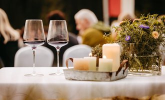 Two wine glasses are placed on a table, surrounded by candles and a small floral decoration. In the background, blurry guests can be seen at a celebration. | © Kleinwalsertal Tourismus eGen | Oliver Farys