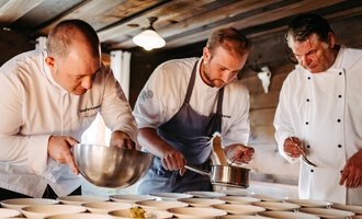Three chefs are working in a wooden hut on the presentation of dishes. They are focusing on their tasks and using various cooking utensils. | © Kleinwalsertal Tourismus eGen | Oliver Farys