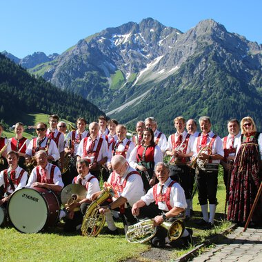 A brass band in traditional attire stands in a picturesque mountain landscape. In the background, there are tall mountains and a blue sky. | © Musikverein d´Hirschegger Kleinwalsertal